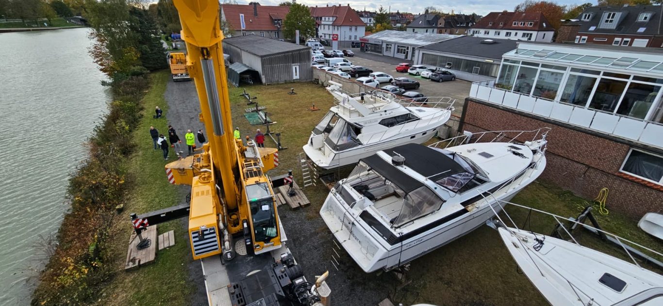 Blick auf zwei Boote, eine Krane und eine Werkstatt am Wasser.