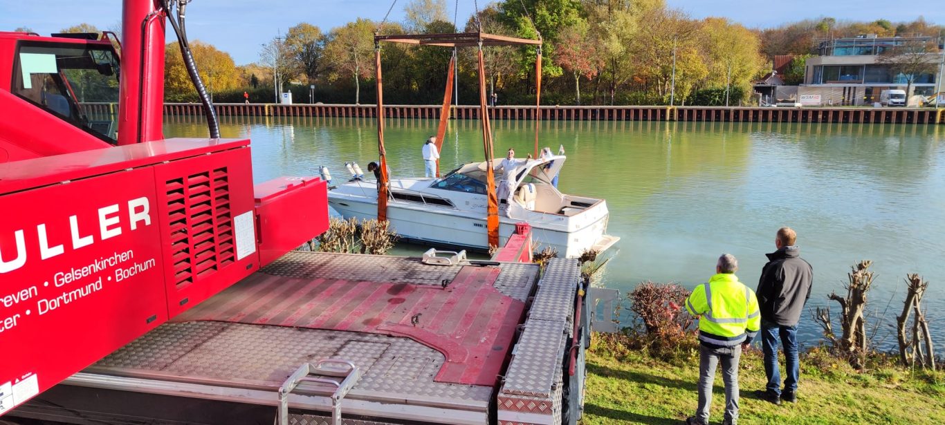 Blick auf einen Hafen mit Booten und zwei Personen am Ufer. Roter Kran im Vordergrund.