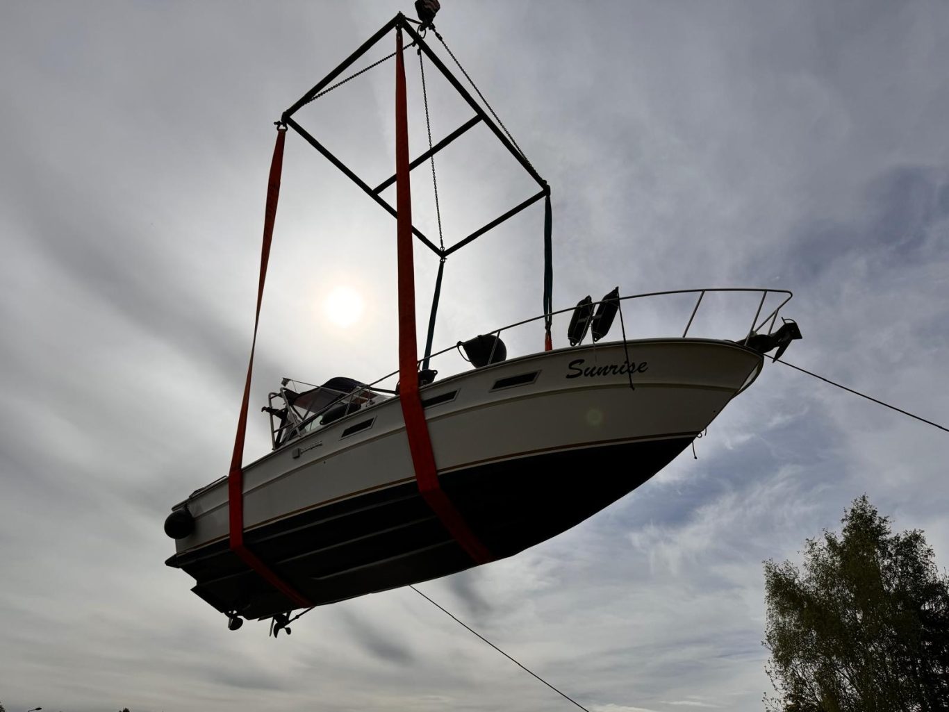Ein Boot wird mit roten Hebebändern über dem Wasser angehoben, Sonnenlicht im Hintergrund.